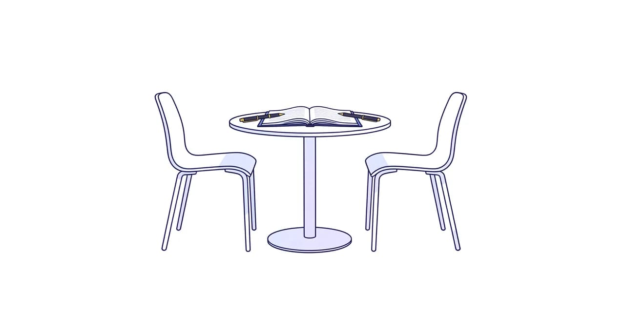 Two chairs drawn up to a small round table, one notebook open flat between them with two pens pointing toward each other.